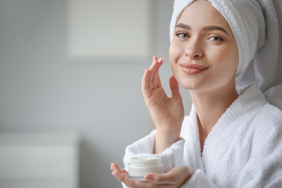 A woman in a bathrobe and hair towel smiling as she applies face cream from a jar